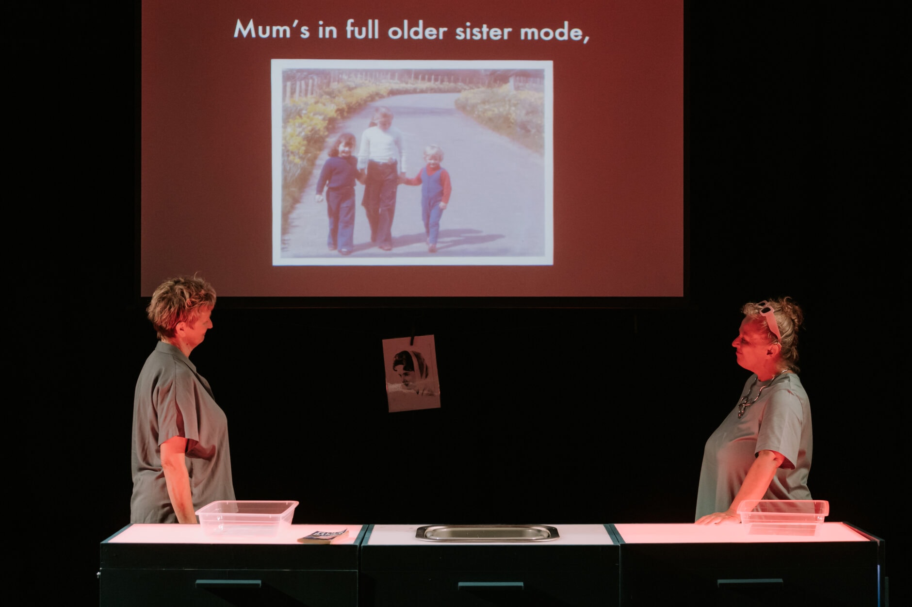 Two women stand at either end of a table. On the screen behind them, there is a photo of two kids and an adult holding hands. The words Mum's in full older sister mode are on the screen.